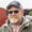 Smiling man with gray beard wearing a cap and sunglasses outdoors, in a jacket and hoodie.