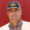 Portrait of a man wearing a brown checkered shirt and a black cap with yellow logo, standing against a red background.