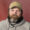 Man with a full beard and mustache wearing a tan cap and gray hoodie, looking at the camera against a red wall background.