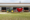 Green tractor pulling a red Jay-Lor feed mixer in a rural barn yard with cows visible in the stalls behind the doors, and a young crop field in the foreground.